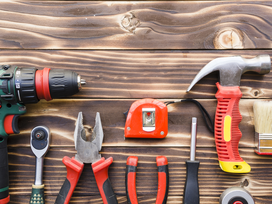 Verschiedene Handwerkszeuge wie Bohrmaschine, Hammer, Zange, Maßband und Schraubendreher auf Holztisch angeordnet.