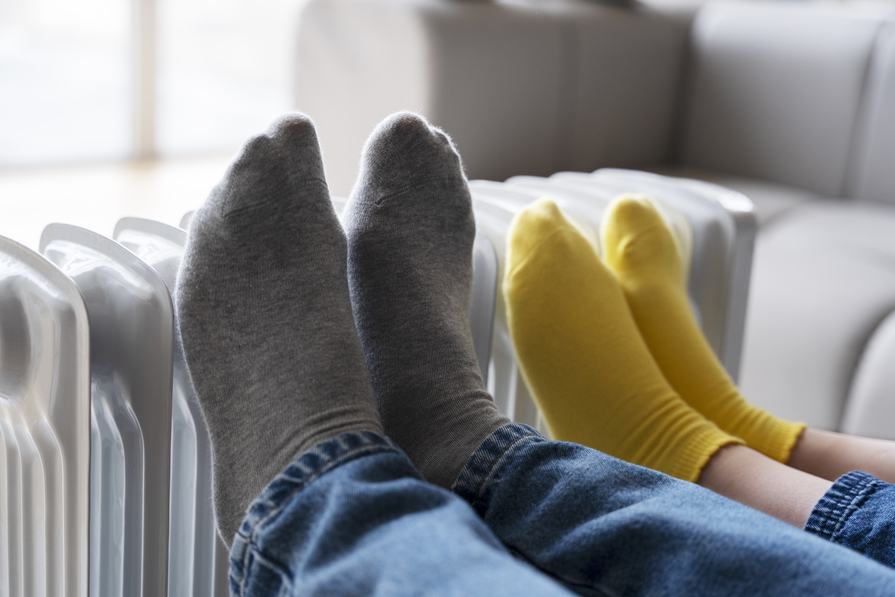 Zwei Personen wärmen ihre Füße in Socken an einem Heizkörper im Wohnraum.