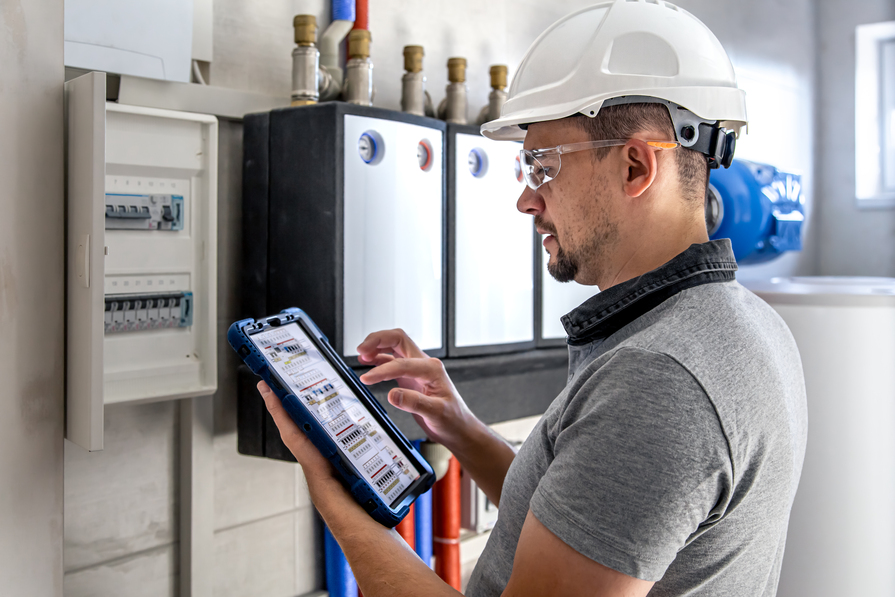 Elektrotechniker mit Schutzhelm prüft elektrische Anlage am Sicherungskasten mithilfe eines Tablets.