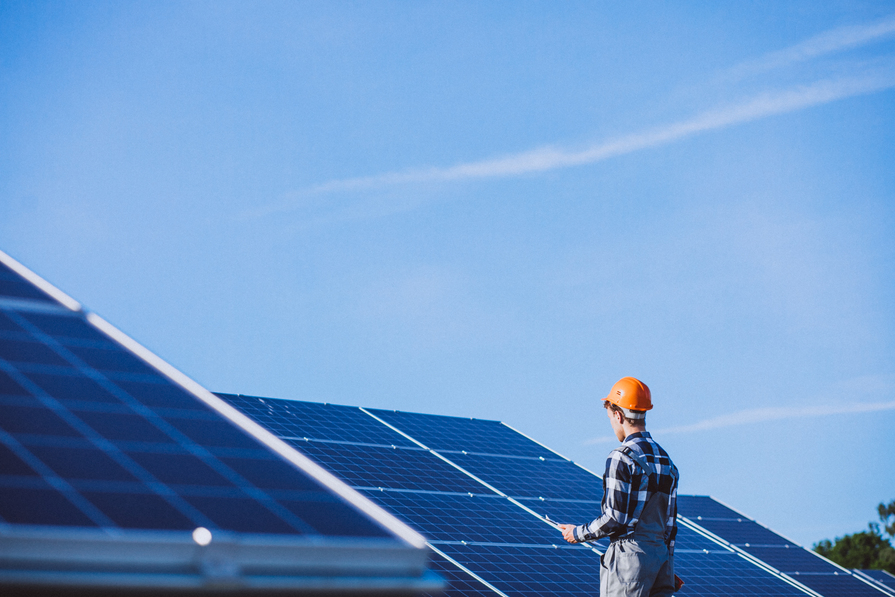 Techniker mit Schutzhelm kontrolliert Photovoltaikanlage auf einem Solarfeld bei klarem Himmel. 