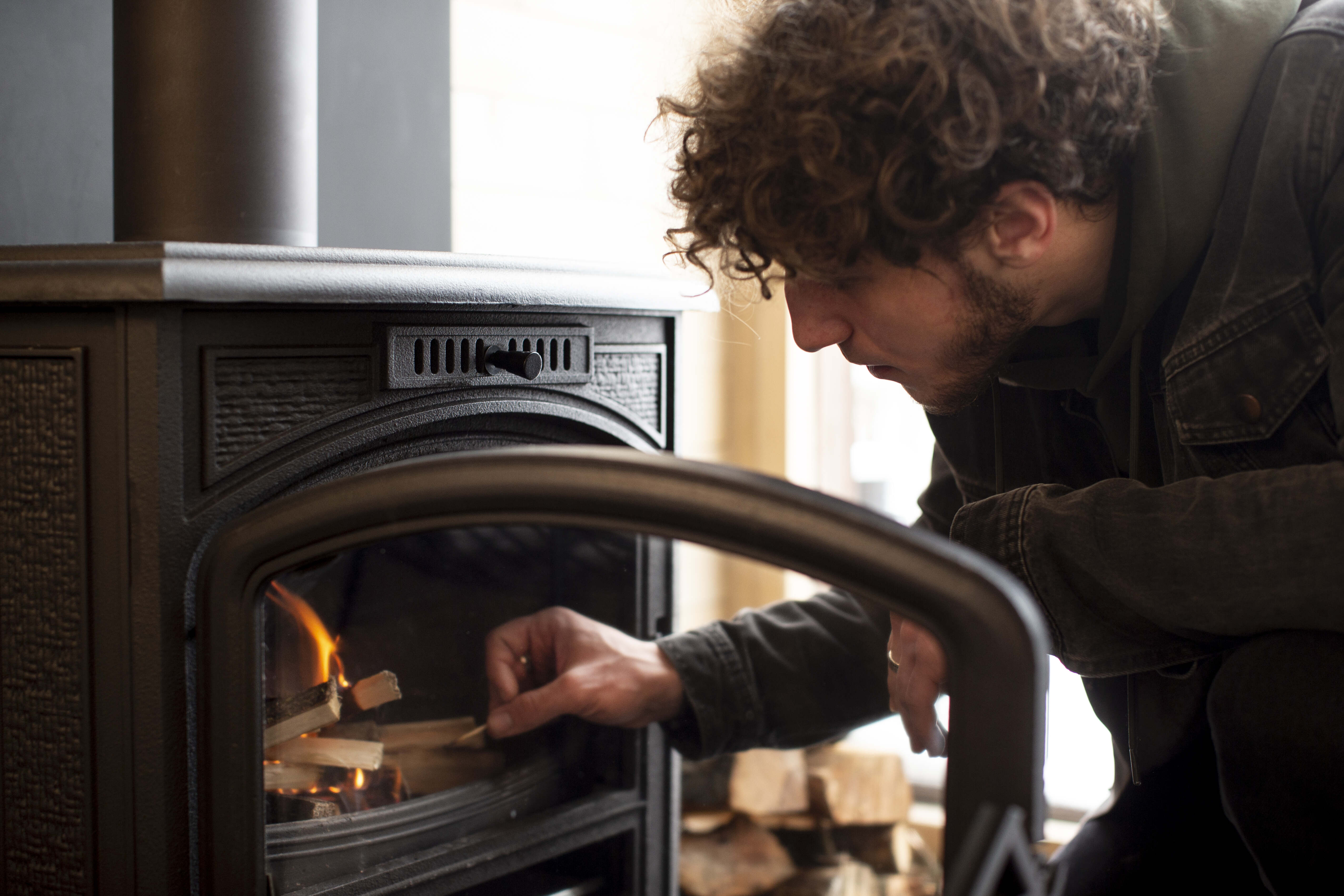 Person legt Holz in einen Kaminofen und entzündet das Feuer in einem gemütlichen Raum.