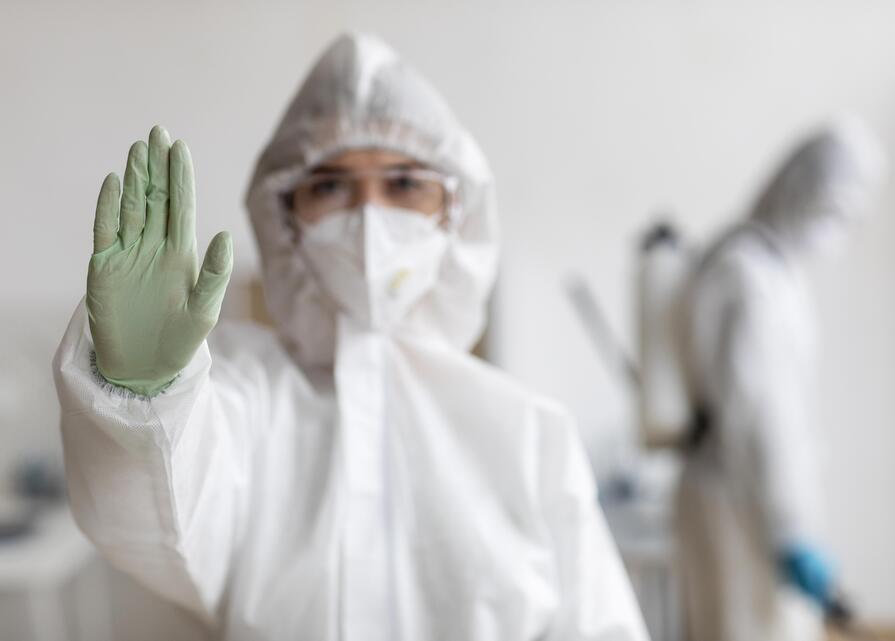  Person in Schutzkleidung mit einer Hand erhoben, als Zeichen zum Stoppen, während im Hintergrund eine weitere Person in Schutzkleidung zu sehen ist.