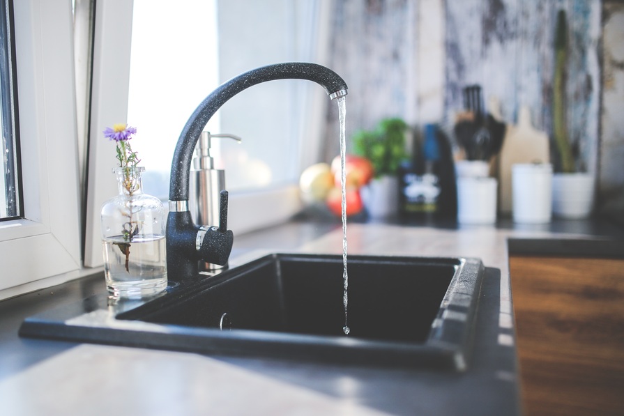Moderne Küchenarmatur mit fließendem Wasser über einem schwarzen Spülbecken, im Hintergrund eine helle Küche mit Arbeitsplatte, Pflanzen und Küchenutensilien.
